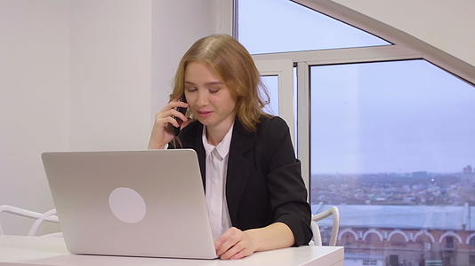 严肃的女商人在办公室电脑前用手机说话视频的预览图