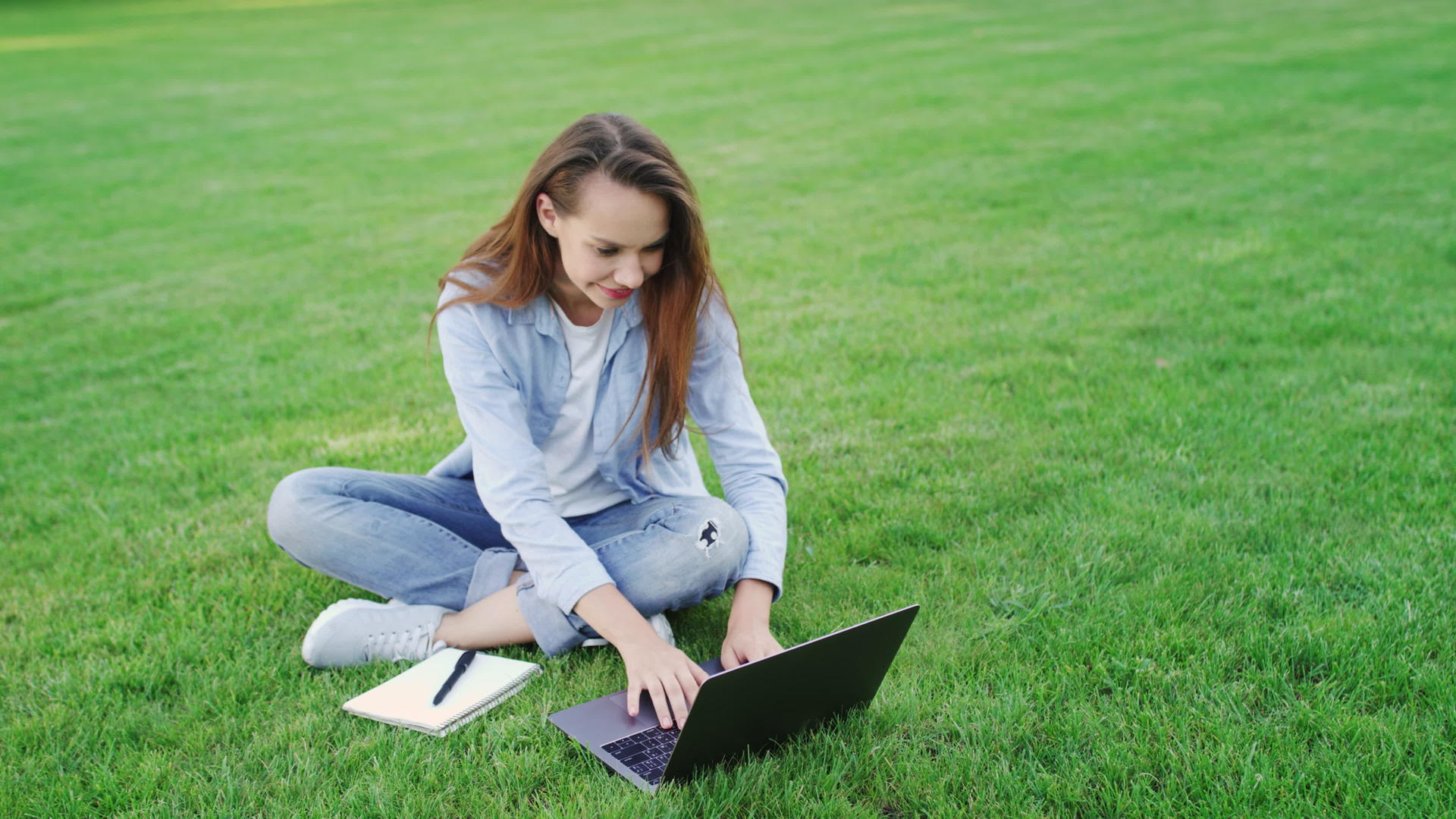 在夏季公园的绿色草坪上工作快乐的女人做笔记本电脑视频的预览图