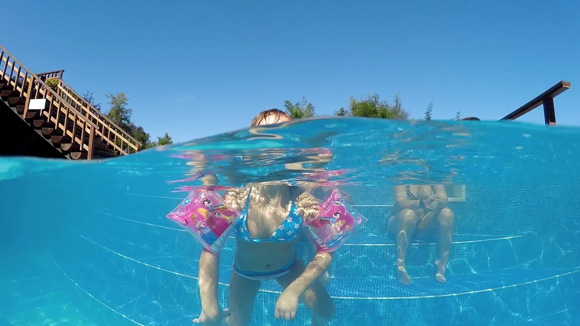 快乐的女孩在漂浮在水面上的游泳池里带着臂带在水面上漂浮飞到相机视频的预览图