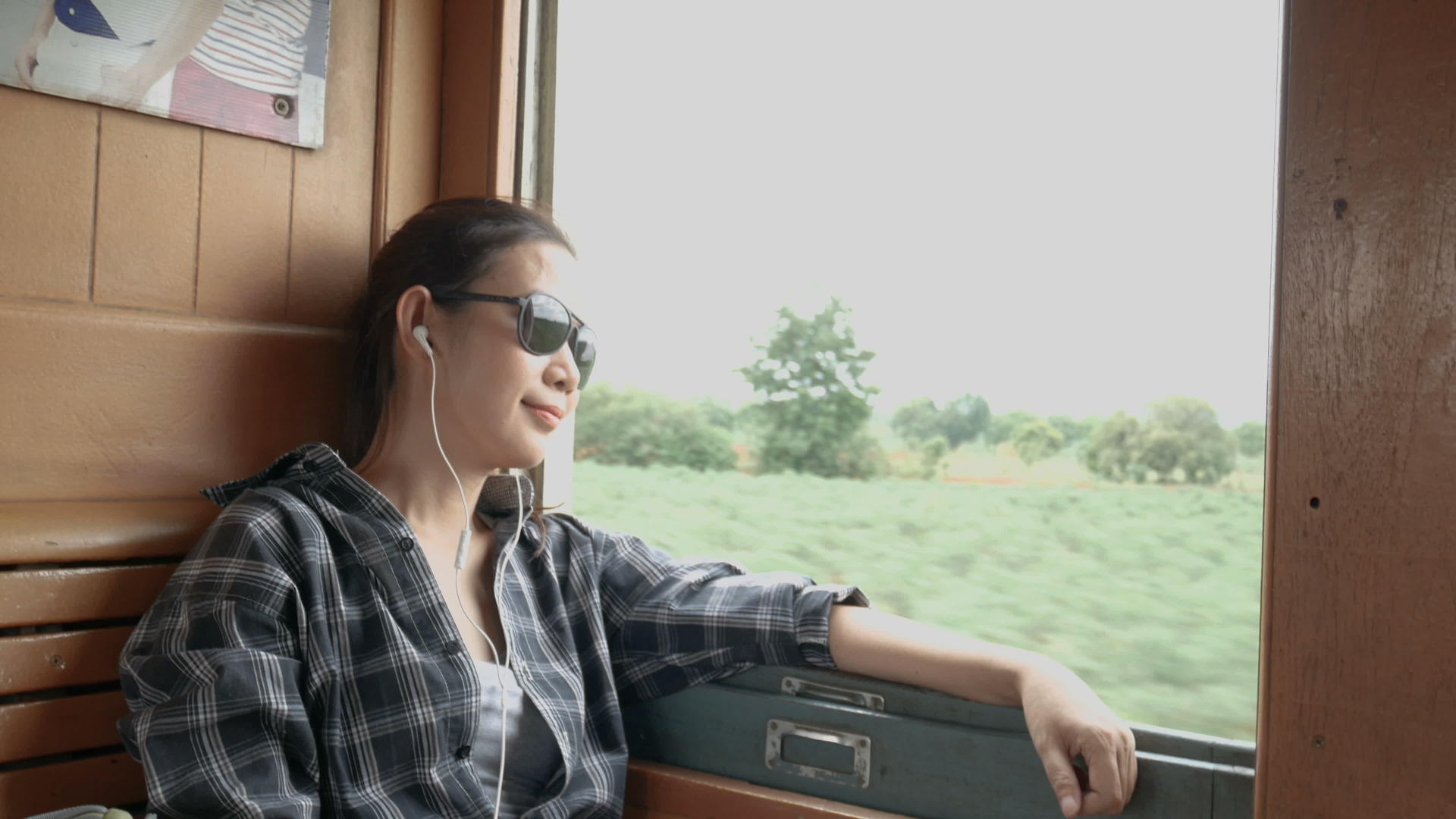 亚洲女性戴着太阳镜用耳机听音乐从曼谷的火车车窗看视频的预览图
