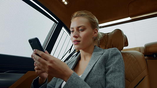 坐在汽车后座上在智能手机上阅读好消息和愉快的白领女性视频的预览图