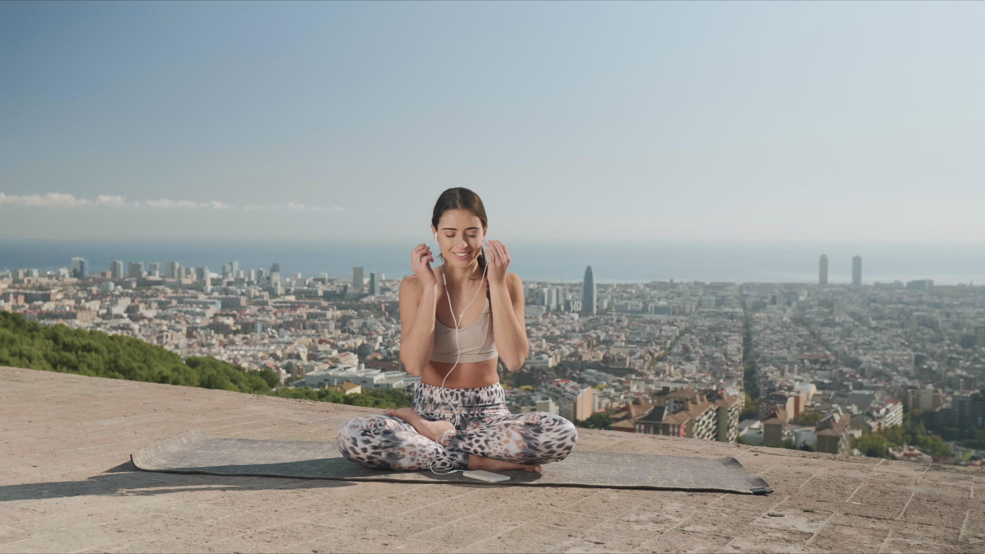 在巴塞罗那的城市背景之前一个女孩盘腿坐在瑜伽垫上听音乐视频的预览图