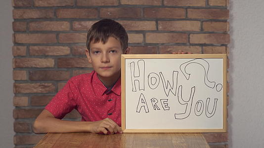 孩子坐在桌子旁拿着一张挂图在背景红砖墙上写着你好吗？视频的预览图