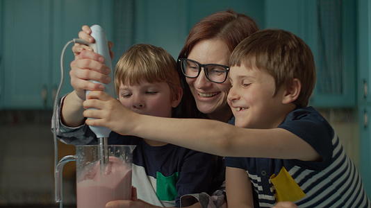 快乐的母亲和两个孩子在家里的蓝色厨房用搅拌机搅拌草莓视频的预览图