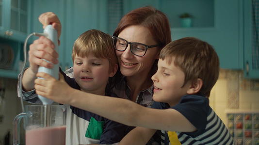 快乐的母亲和两个孩子在家里的蓝色厨房里用搅拌机做果汁视频的预览图