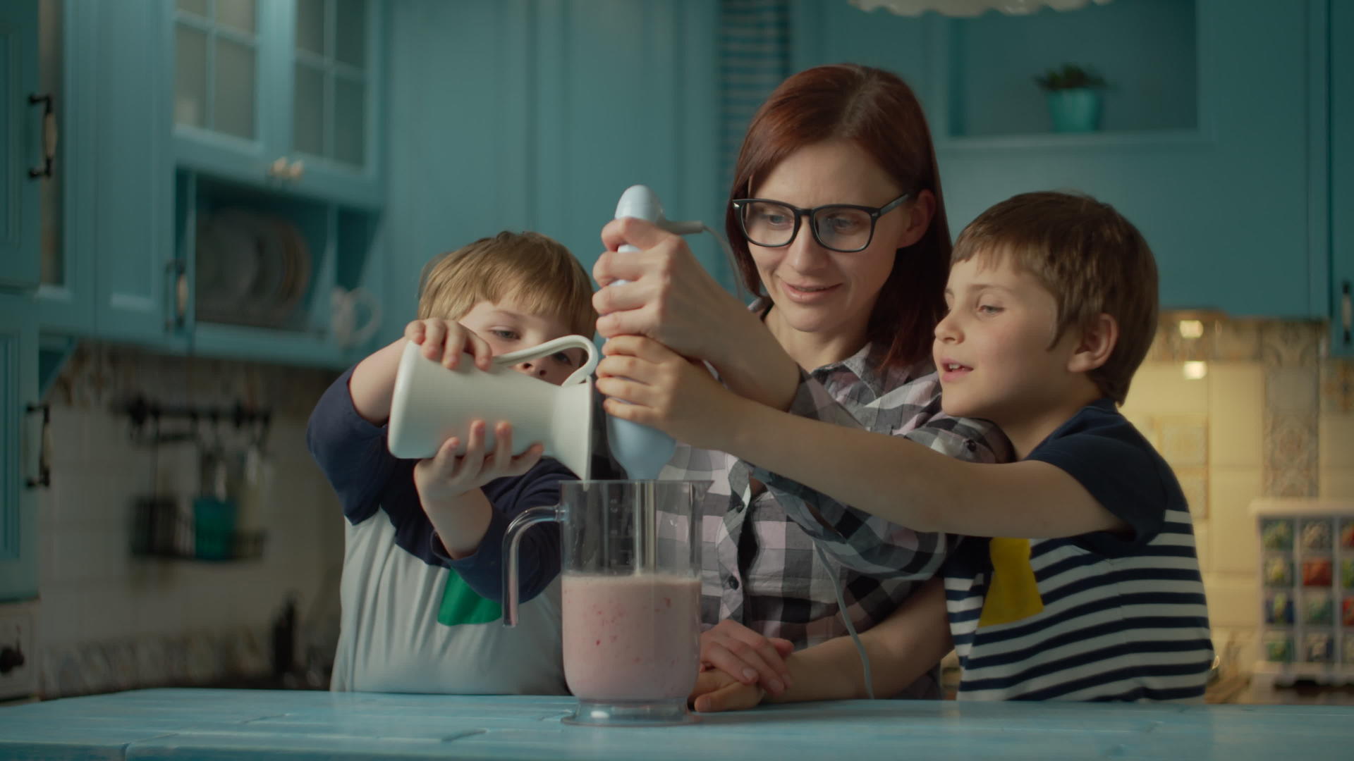 快乐的母亲和两个孩子用搅拌机在家里的蓝色厨房里煮草莓泥视频的预览图