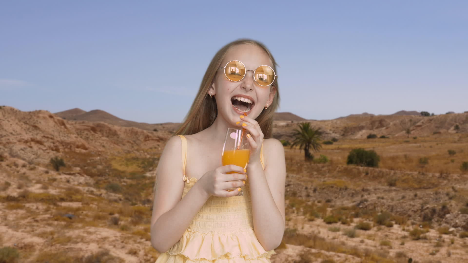 兴奋的女孩在沙漠和山脉的背景上用橙汁玻璃笑着视频的预览图